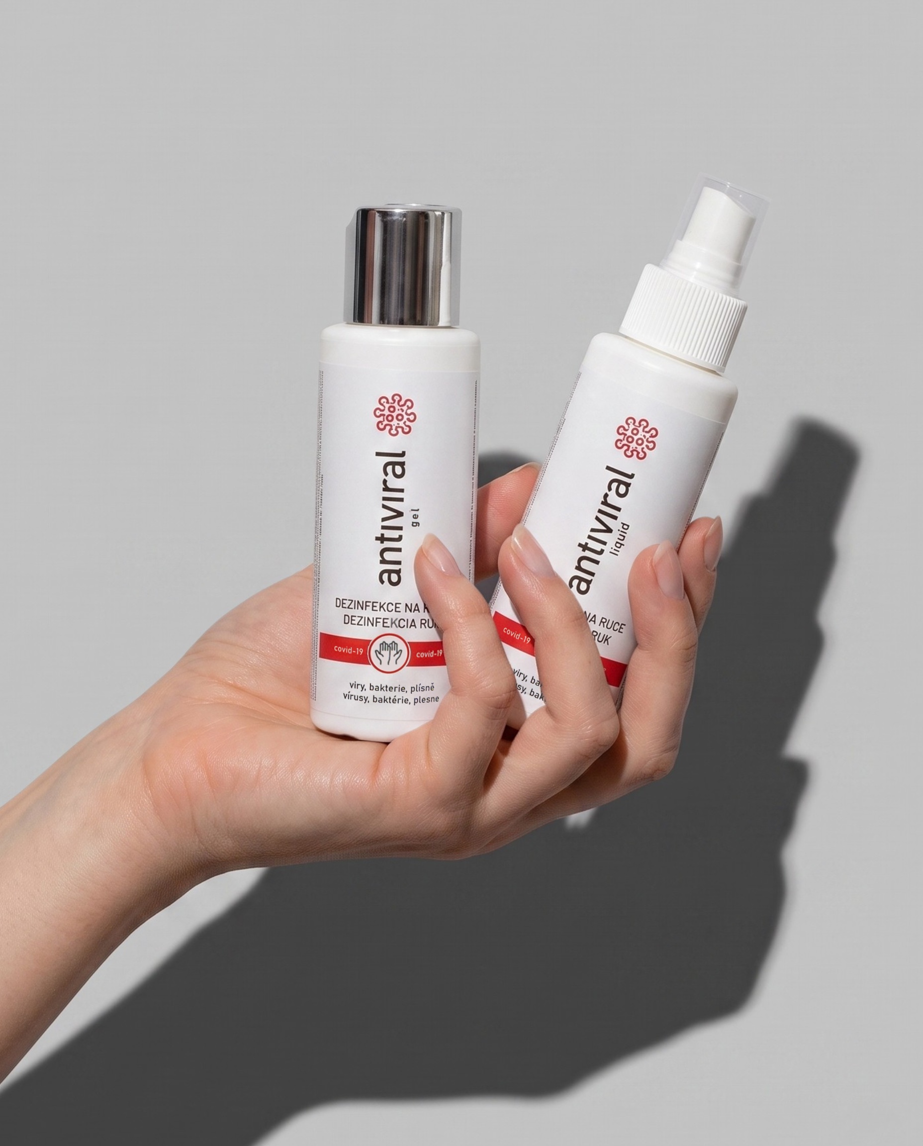 Ruka drží vedle sebe lahvičku ANTIVIRAL gel a liquid, vržený stín na světle šedé ploše