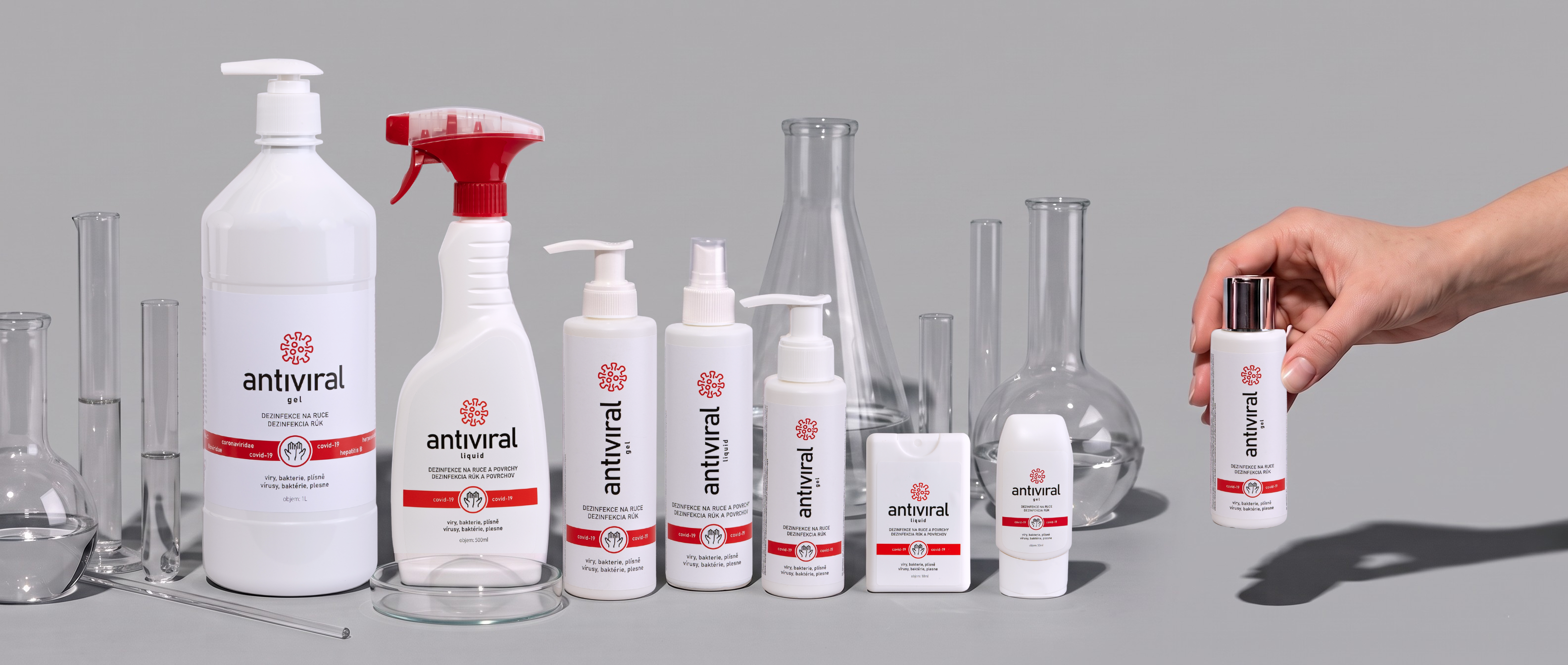 Celá řada balení ANTIVIRAL gel a liquid mezi laboratorním sklem na šedém pozadí, ruka z pravého okraje zvedá kapesní balení gelu